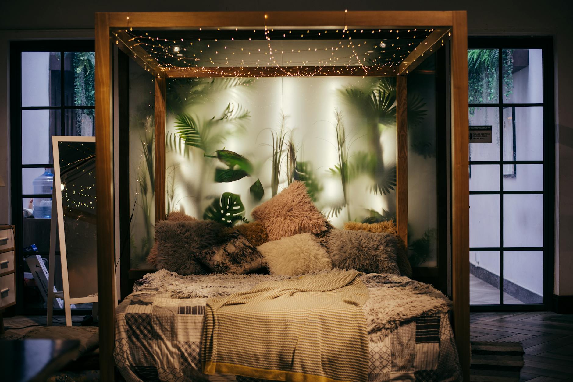 a super cosy (with slightly too furry pillows) bed surrounded by a bedframe with fairy lights and a milky glass wall in the back with green palm trees behind it